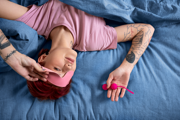 woman laying on bed with eye mask on and vibrator in hand.